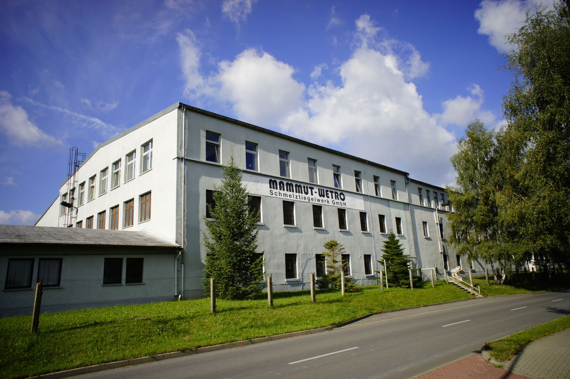 Das weiße Firmengebäude mit dunklem Dach unter blauem Himmel mit Wolken von Mammut Wetro.